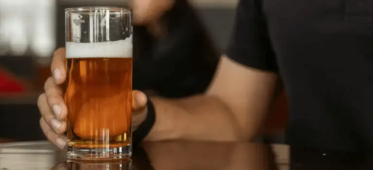 Person holding a glass of beer on a bar counter, with another patron blurred in the background
