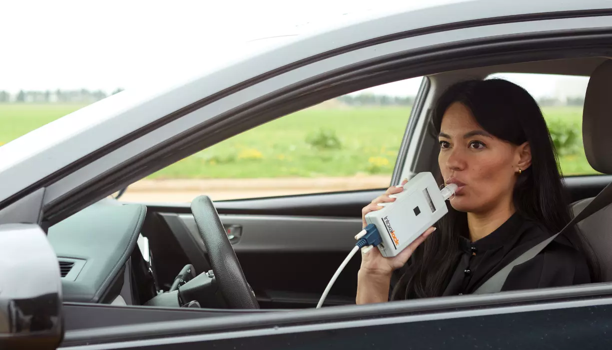 A woman sitting in the driver’s seat of a car blowing into the mouthpiece of an Intoxalock ignition interlock device