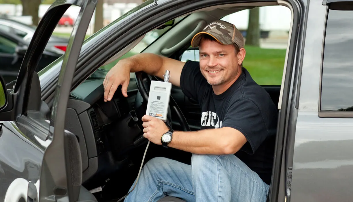 A man sitting in the driver's seat of a car, holding an ignition interlock device, ready to perform a breathalyzer test before starting the vehicle.