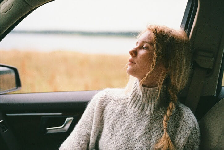 A woman is seated in the passenger side of a car, looking out the window at a serene field, reflecting on her thoughts or mental health in a moment of tranquility.