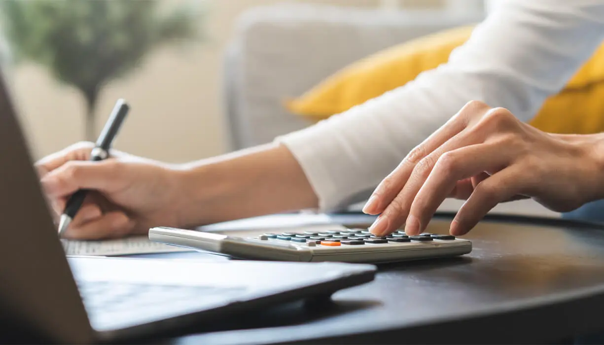 A person calculating finances on a calculator beside a laptop, with a notebook and pen on the table